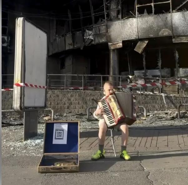 На Лукʼянівці дитина грає на акордеоні біля зруйнованого росіянами торгового центру